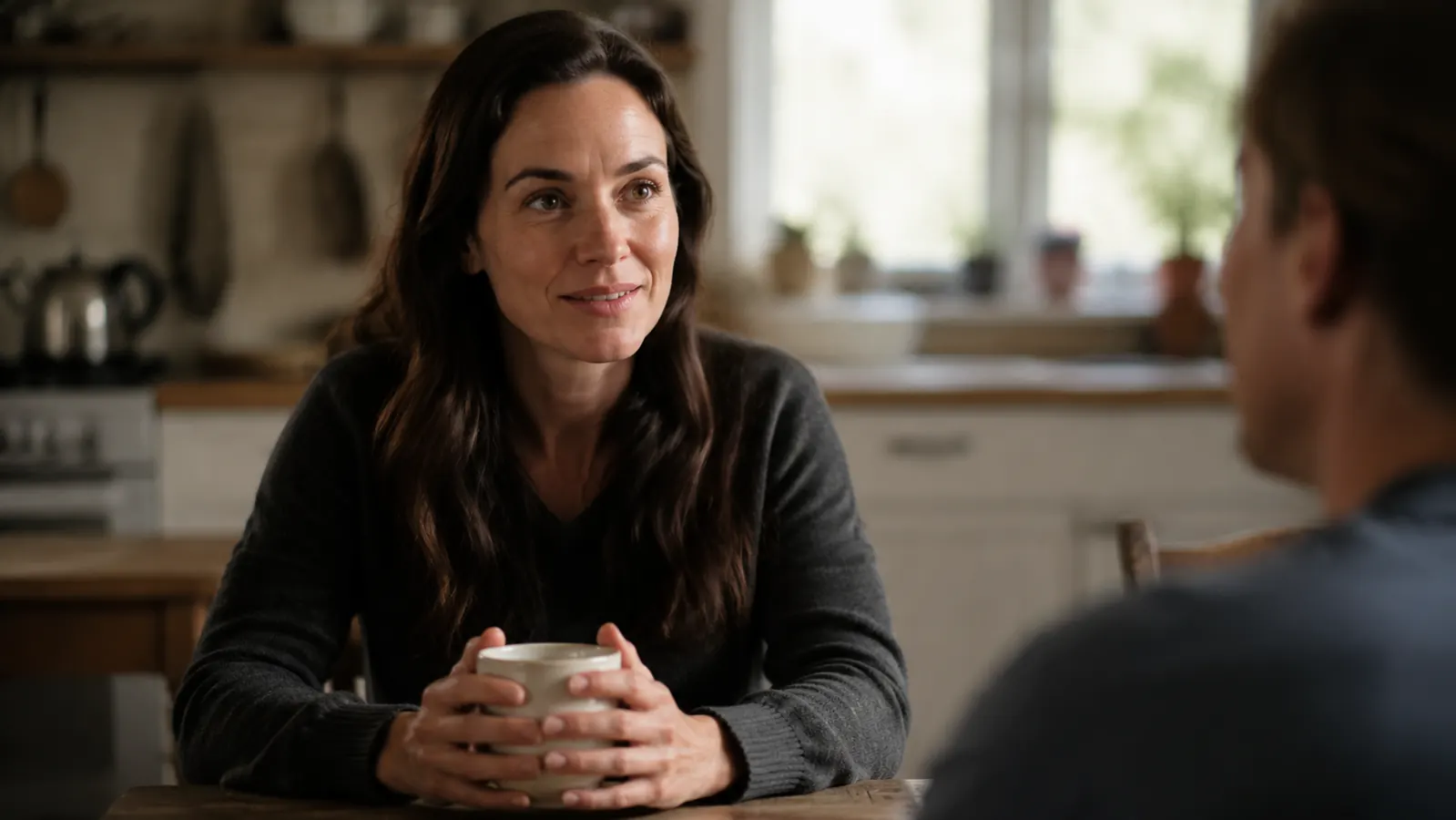 Person sitting at a kitchen table with a cup of tea, relaxed expression, morning light coming through the window