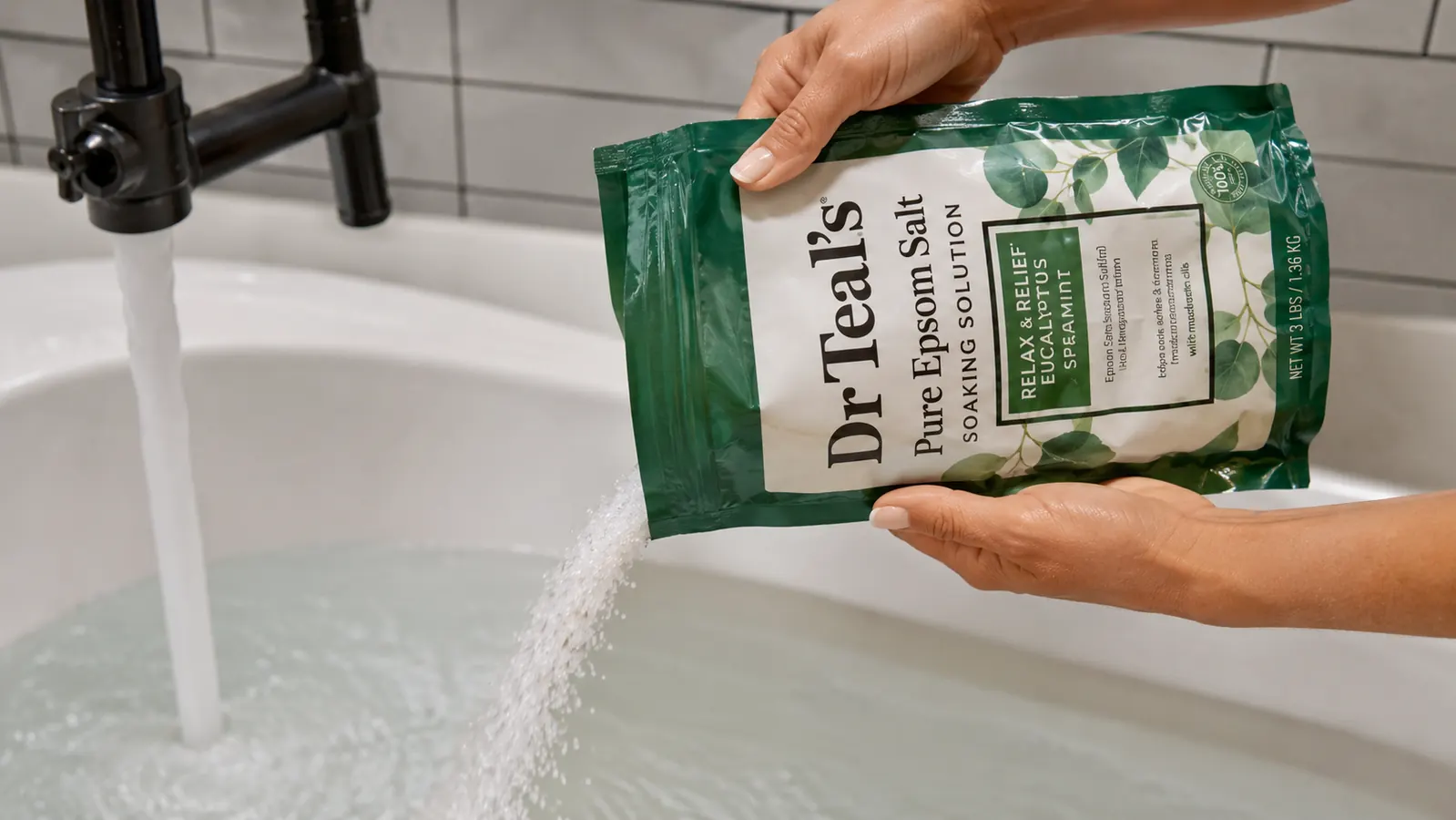 Hand pouring epsom salt crystals from a green Dr Teal's bag into a running bathtub
